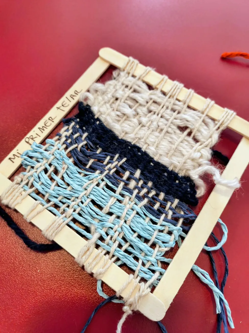 Telar en miniatura con la inscripción Mi primer telar, tejido en lana celeste, azul y natural