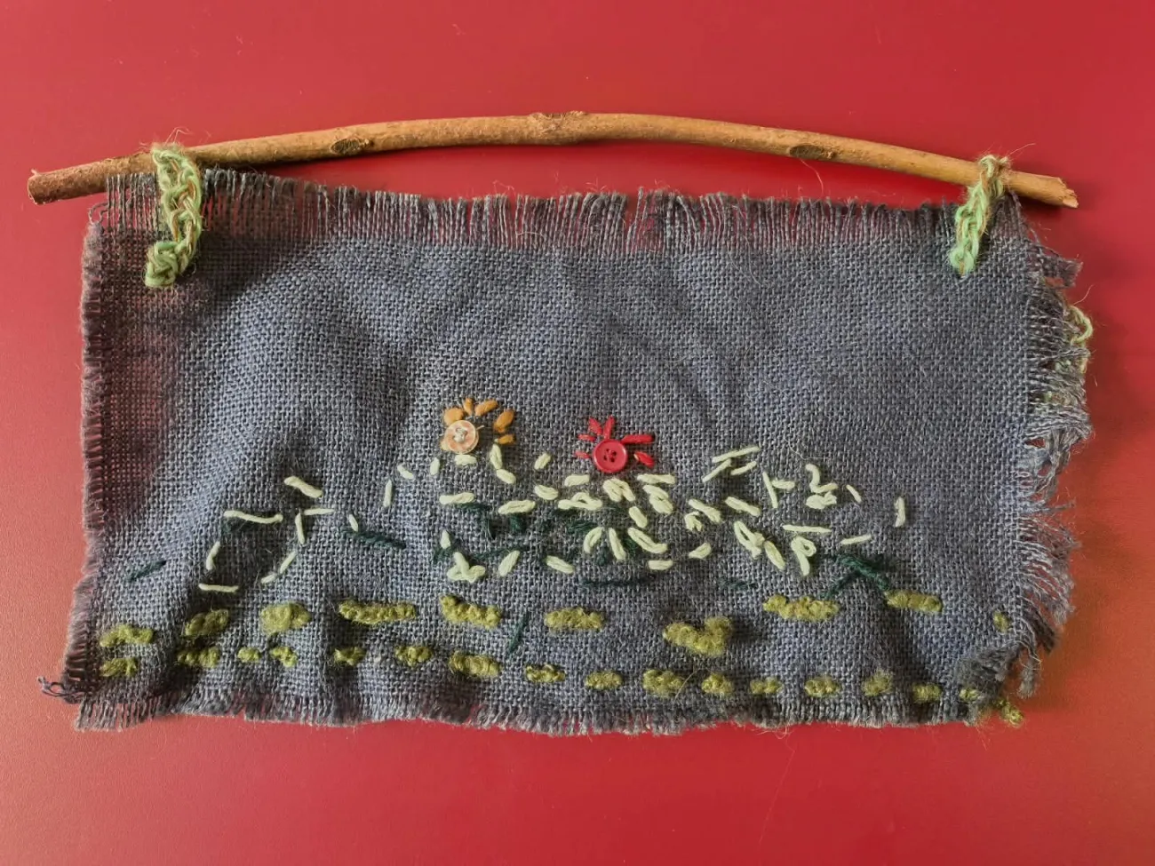 Tapiz bordado a mano sobre arpillera con flores, colgado de una rama